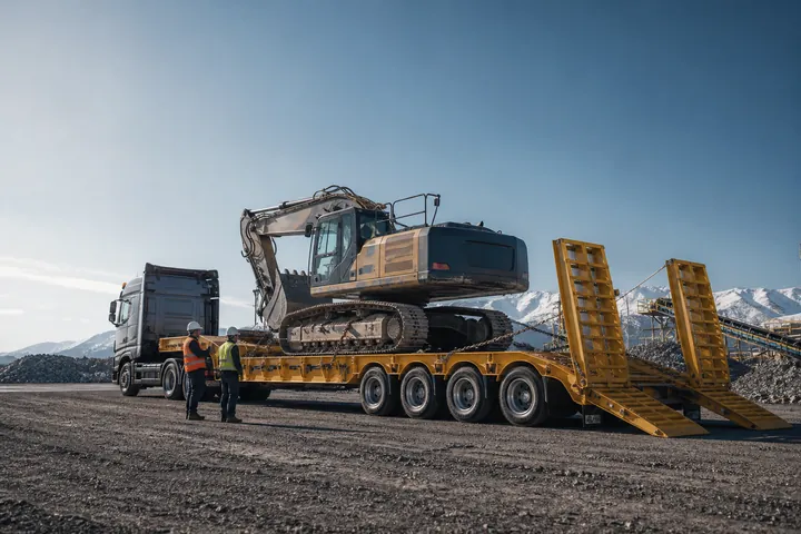 Excavadora sobre una cama baja con rampas en una zona de trabajo.